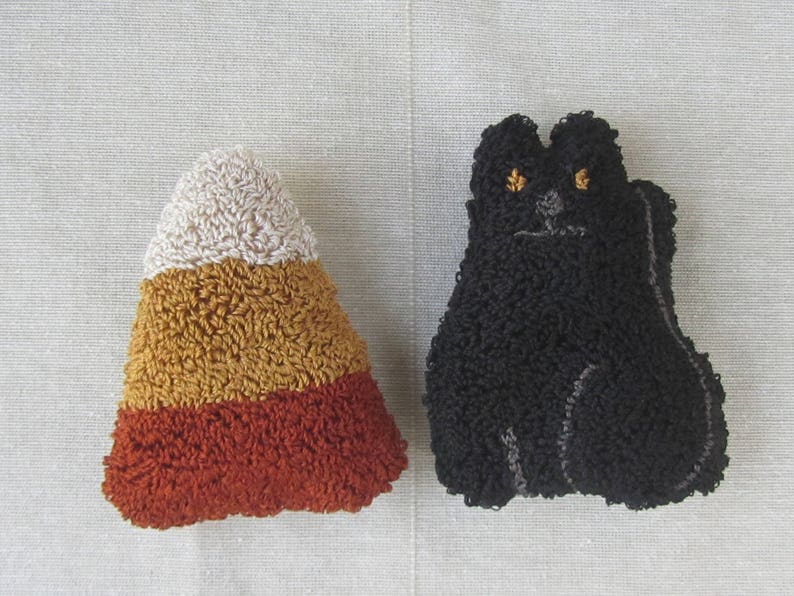 May include: Two handmade Halloween decorations. One is a candy corn in white, yellow, and orange. The other is a black cat with yellow eyes. Both are made with a textured, looped yarn.