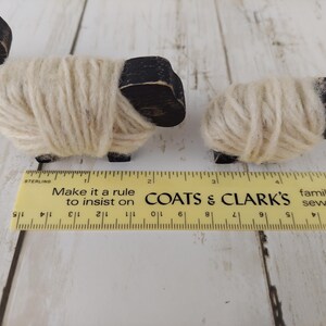 May include: Three small wooden sheep figurines with black heads and legs, and wrapped in cream-colored yarn. A yellow ruler with black markings and text is in the foreground. The text on the ruler says "Make it a rule to insist on COATS & CLARK'S".