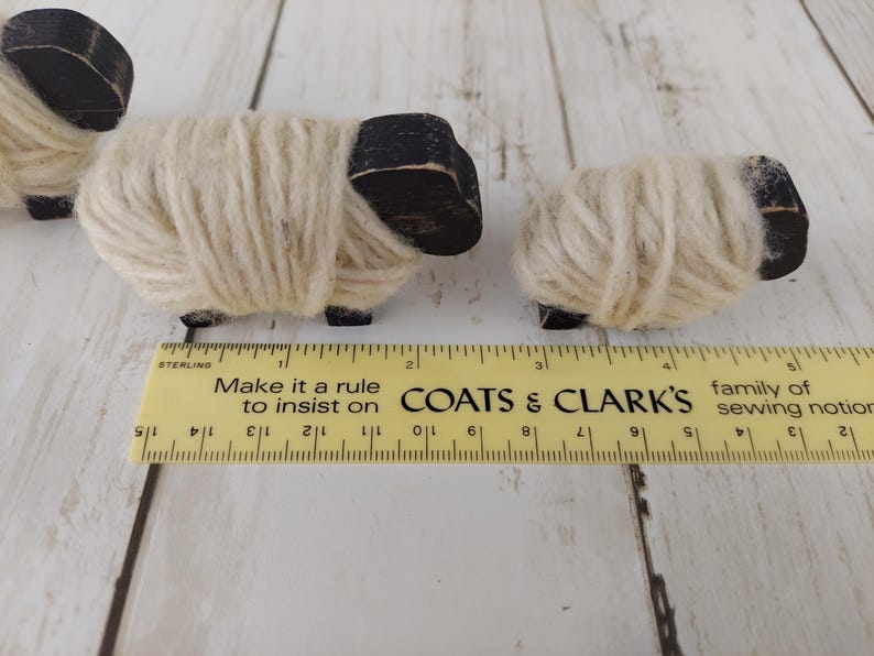May include: Three small wooden sheep figurines with black heads and legs, and wrapped in cream-colored yarn. A yellow ruler with black markings and text is in the foreground. The text on the ruler says "Make it a rule to insist on COATS & CLARK'S".