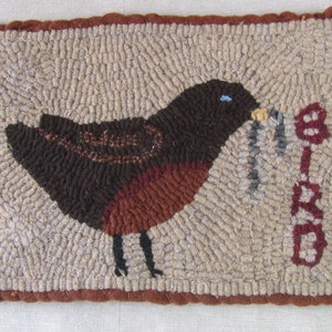 May include: Hand-hooked rug featuring a brown bird with a red breast, holding a worm in its beak. The word "BIRD" is stitched in red on the right side. The rug has a beige background and a brown border.