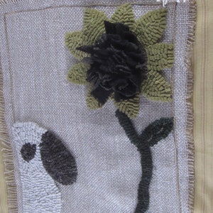 May include: Handmade textile art piece featuring a sunflower with a black center and green petals, a dark green stem, and a white and gray shape on a burlap background. The edges are frayed, adding a rustic touch to the artwork.