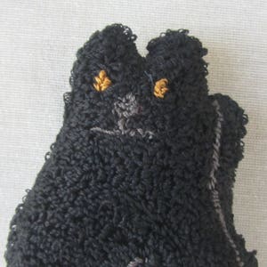 May include: A close-up of a black cat-shaped textile item with a textured, looped surface. The cat has embroidered golden eyes and a grey nose and mouth. The item is likely a decorative piece or a small toy.
