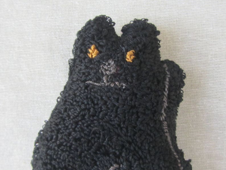 May include: A close-up of a black cat-shaped textile item with a textured, looped surface. The cat has embroidered golden eyes and a grey nose and mouth. The item is likely a decorative piece or a small toy.