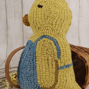 May include: A handmade yellow duck-shaped pillow with a blue and brown design. The duck has a textured, looped surface and a small brown beak. The pillow is placed near a woven basket and white flowers.