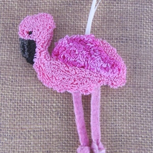 Könnte beinhalten: Ein rosa Flamingo-Ornament mit strukturiertem Körper, schwarzem Schnabel und langen rosa Beinen. Der Flamingo hat eine weiße Schnur zum Aufhängen. Das Ornament besteht aus einem weichen, plüschigen Material.