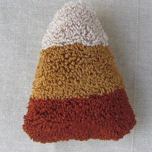 May include: A crocheted candy corn decoration. The triangular shape is made up of three horizontal stripes: cream at the top, golden yellow in the middle, and a deep orange at the bottom. The stitches create a textured surface.