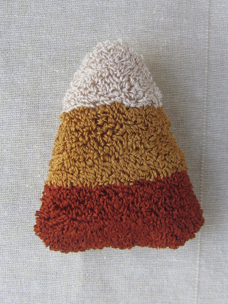 May include: A crocheted candy corn decoration. The triangular shape is made up of three horizontal stripes: cream at the top, golden yellow in the middle, and a deep orange at the bottom. The stitches create a textured surface.