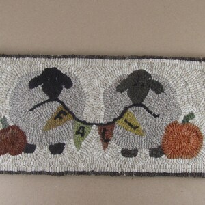 May include: A rectangular, hand-stitched rug featuring two black sheep flanking a banner that spells "FALL". The sheep are set against a light gray background, with a pumpkin on each side. The rug has a dark brown border.