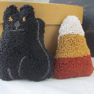 May include: A black cat-shaped plush toy with gold eyes and a grey embroidered face, beside a candy corn-shaped plush toy. The candy corn plush has white, yellow, and orange sections. Both are made of textured fabric.