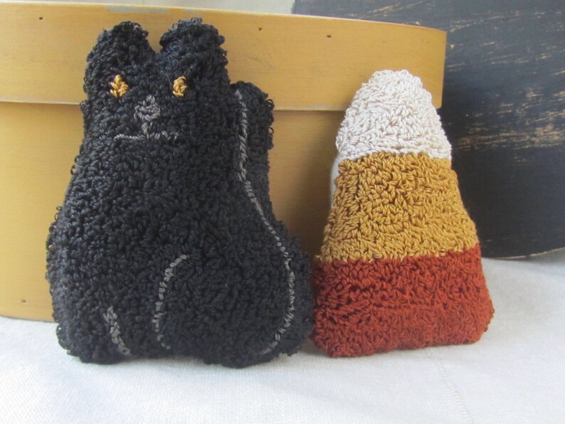 May include: A black cat-shaped plush toy with gold eyes and a grey embroidered face, beside a candy corn-shaped plush toy. The candy corn plush has white, yellow, and orange sections. Both are made of textured fabric.