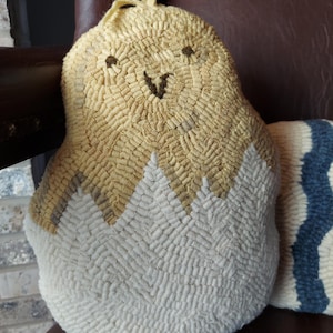 May include: A handmade, textured pillow in the shape of a pear. The top is light yellow with brown embroidered eyes and a smile. The bottom half is white. The pillow is resting on a brown leather chair.