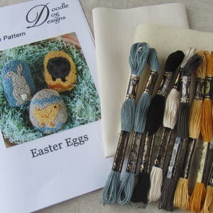 May include: A punch needle pattern kit with the title "Easter Eggs" featuring a bunny, sheep, and chick design. The kit includes embroidery floss in various colors, fabric, and a pattern sheet. The floss colors include blue, black, and tan.