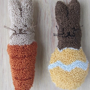 May include: Two handmade Easter bunny decorations. One bunny is beige and orange, the other is brown and yellow with a blue stripe. Both have stitched whiskers and are made of a textured material, possibly terry cloth.