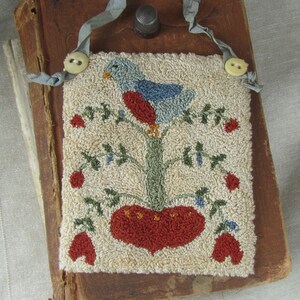 Puede incluir: Un adorno de pared rectangular cosido a mano con un pájaro azul y rojo posado en un árbol verde con un corazón y flores rojas. Se adjunta a un libro antiguo con una cinta y un botón. El fondo es de tela neutra y texturizada.