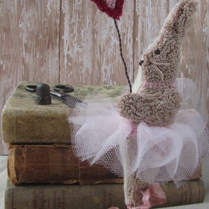 May include: A handmade bunny doll with a pink tutu and a heart-shaped balloon. The bunny is light brown with pink accents, and the tutu is made of white tulle. The heart balloon is red and pink. The doll is placed on top of old books.