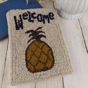May include: A rectangular, beige rug with a pineapple design and the word "welcome" in navy blue. The pineapple is brown with a dark brown crown. The rug is made of a textured material, and is on a white wooden surface.