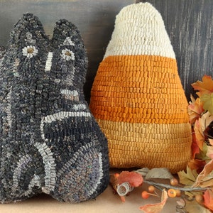 Puede incluir: Cojines hechos a mano con temática de Halloween. Un cojín tiene forma de gato negro con detalles blancos, y el otro tiene forma de caramelo de maíz con secciones blancas, naranjas y amarillas. Los cojines están hechos de un material texturizado.