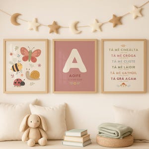 May include: Three framed wall art prints featuring a butterfly, bee, snail, and ladybug on the left, a large letter A with the name Aoife in the centre, and Irish text with English translations on the right, all displayed above a bench with a plush rabbit.