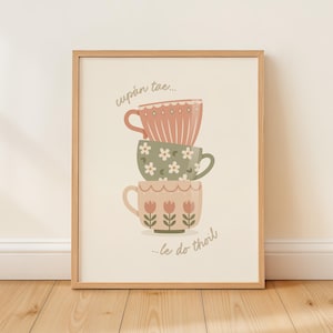 May include: A framed wall art print featuring a stack of three illustrated tea cups in muted pink and sage green tones. The top cup is pink with vertical lines, the middle is green with white flowers, and the bottom is pink with tulip motifs. Text reads cupán tae... le do thoil.