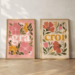 Op de afbeelding: Twee ingelijste rechthoekige wandposters met botanische illustraties. De linker poster heeft een roze achtergrond met de witte tekst "grá" omringd door roze, gele en groene bloemen. De rechter poster heeft een crèmekleurige achtergrond met de gele tekst "croí" omringd door rode, roze en groene bloemmotieven.