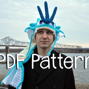 May include: A person wearing a blue and white crocheted dragon hat with a long tail. The hat has a pointed top and a wide brim. The person is standing in front of a bridge and a body of water. The text "PDF Pattern" is written in white letters on a black background.