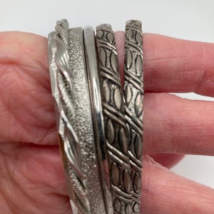 May include: A stack of silver bangles with varying textures and designs. The bangles include a twisted rope pattern, a textured band, a smooth band, and two bands with an embossed leaf pattern.