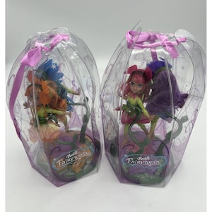 May include: Two Barbie Fairytopia dolls in clear plastic packaging. One doll has orange and blue wings, the other has pink and purple wings. The packaging is decorated with purple accents and a pink ribbon.