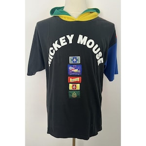 May include: Black short-sleeved t-shirt with a yellow, green, and blue color block design. The shirt has a Mickey Mouse logo with the text "Mickey Mouse" in white. The shirt has a hood and features several colorful patches.