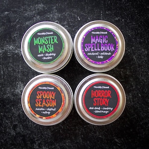 May include: Four round silver metal tins with labels. Top left: green label reads Monster Mash, apple + blueberry + bourbon. Top right: purple label reads Magic Spellbook, parchment + patchouli + herbs. Bottom left: orange label reads Spooky Season, pumpkin + chestnut + nutmeg. Bottom right: red label reads Horror Story, dark woods + cranberry + blood orange.