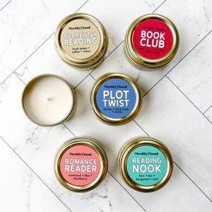 Könnte beinhalten: Eine Auswahl an Duftkerzen in goldenen Dosen. Jede Kerze hat ein Etikett mit einem einzigartigen Namen und einer Duftbeschreibung, darunter "Currently Reading", "Book Club", "Plot Twist", "Romance Reader" und "Reading Nook".