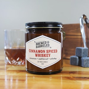 An amber glass jar labeled "BREWED & BARRELED CINNAMON SPICED WHISKEY" sits on a wooden surface, accompanied by a glass of amber-colored liquid and a set of gray cubes.