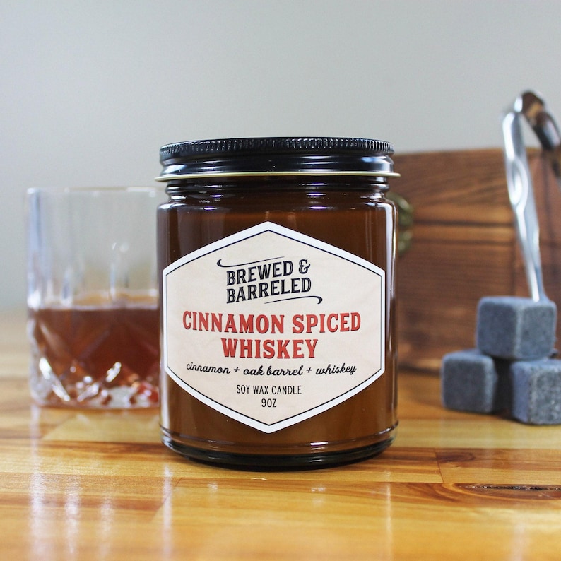 An amber glass jar labeled "BREWED & BARRELED CINNAMON SPICED WHISKEY" sits on a wooden surface, accompanied by a glass of amber-colored liquid and a set of gray cubes.