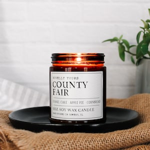 May include: A brown glass jar candle with a white label that reads "Novelly Yours County Fair Funnel Cake Apple Pie Cornbread 9 oz Soy Wax Candle Hand-Poured in Oswego, IL"