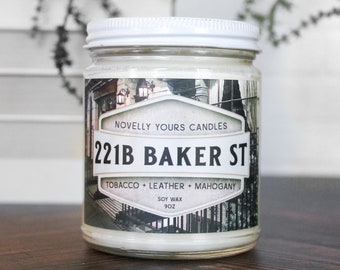 May include: A glass jar candle with a white lid and label featuring the text "221B BAKER ST". The label also includes "TOBACCO + LEATHER + MAHOGANY" and "SOY WAX 9OZ". The candle sits on a dark wooden surface.