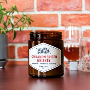 An amber glass jar labeled "Brewed & Barreled Cinnamon Spiced Whiskey" sits on a wooden surface, with a glass of amber liquid and a plant in the background.