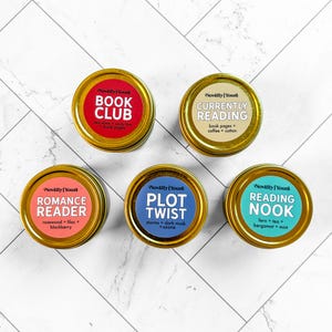 May include: Five round gold-coloured candle tins with labels reading Novelly Yours. The labels are red, beige, pink, blue, and teal, featuring the text Book Club, Currently Reading, Romance Reader, Plot Twist, and Reading Nook with scent notes.