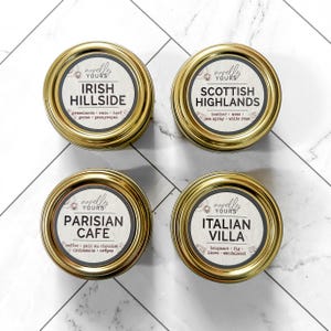Könnte beinhalten: Vier runde, goldfarbene Kerzendosen mit Etiketten. Die Etiketten lauten "Irish Hillside", "Scottish Highlands", "Parisian Cafe" und "Italian Villa". Jedes Etikett listet Duftnoten auf. Die Dosen sind auf einer weiß-grau gemusterten Oberfläche angeordnet.