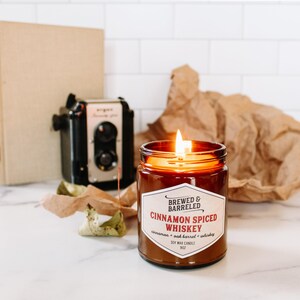 A lit cinnamon-spiced whiskey candle in the foreground, with a vintage camera and brown paper in the background, creating a cozy and nostalgic atmosphere.