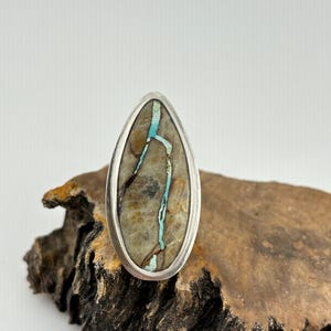 May include: A teardrop-shaped ring with a silver bezel. The ring features a stone with a tan and brown pattern, accented by a turquoise-colored vein. The ring is displayed on a piece of textured wood, showcasing the natural stone's unique design.