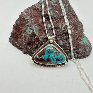 May include: A silver pendant necklace featuring a triangular pendant with a turquoise and brown stone. The pendant is suspended from a delicate silver chain. The stone has a unique pattern and texture.