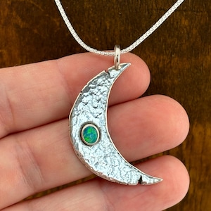 May include: A silver crescent moon pendant with a textured surface and a small, round, green gemstone set in the center. The pendant hangs from a delicate silver chain. The background is a dark brown.