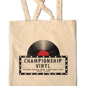May include: A beige tote bag with a black and white illustration of a vinyl record and a red label. The text on the bag reads "Championship Vinyl" and includes the address "1500 North Milwaukee Avenue & North Honore Street, Chicago, IL 60622".