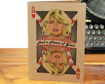 May include: A greeting card featuring a pop art style illustration of a woman with blonde hair, framed as a playing card. The card reads "Had a Heart of Glass" and includes red hearts. The card is on a wooden surface.