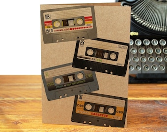 May include: A brown card with four vintage cassette tapes on it. The tapes are different colors and have different labels. The card is sitting on a wooden table next to a vintage typewriter.