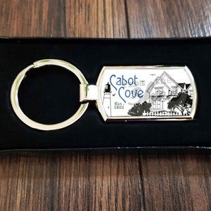 May include: Silver keyring with a black and white image of a lighthouse and house with the text "Cabot Cove Est. 1885".