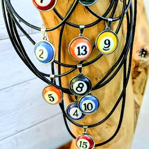 Peut inclure: Plusieurs colliers à cordon noir, chacun orné d'un pendentif en forme de boule de billard. Les pendentifs affichent les numéros 2, 4, 5, 7, 8, 9, 10, 11, 13, 14 et 15, avec leurs couleurs et motifs correspondants. Les colliers sont exposés sur un présentoir en bois.