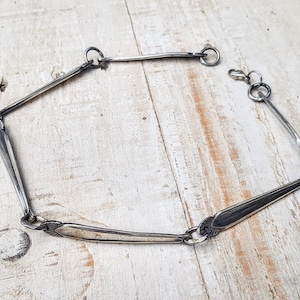 May include: A silver necklace made from repurposed silverware. The necklace is made of several spoon handles connected together with silver rings. The necklace is lying on a white wooden surface.