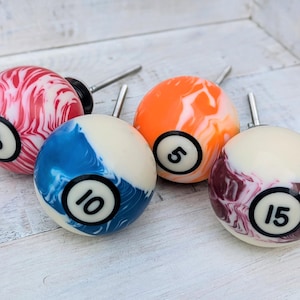 May include: Four cabinet knobs designed as billiard balls. The knobs are in various colours: red and white, orange and white, blue and white, and burgundy and white. Each has a number from a billiard ball, 3, 5, 10, and 15.