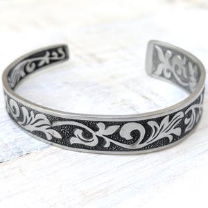 May include: A silver-toned metal cuff bracelet featuring a continuous embossed floral scroll pattern. The design consists of raised, polished silver-colored leaf motifs set against a recessed, darkened, and textured background along the entire band.