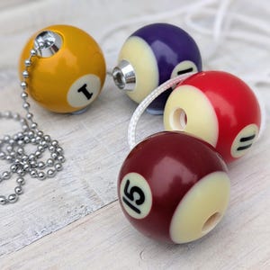 May include: Four billiard ball-shaped light pulls in yellow, purple, red, and maroon. Each ball has a number and a cream-coloured section. The yellow ball has a silver chain, while the others have white cords. The pulls are on a light wood surface.